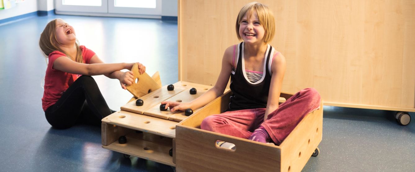 Zwei lachende Mädchen spielen mit einem selbstgebauten Holzwagen, in dem eine von ihnen sitzt