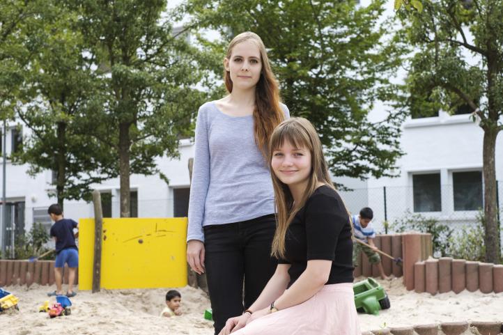 Zwei junge Frauen sitzen auf einem Spielplatz mit spielenden Kindern im Hintergrund