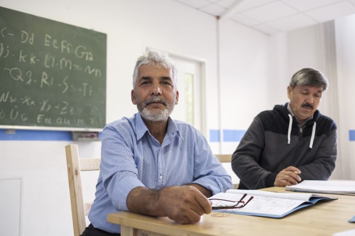 Bild, auf dem zwei Männer, geflüchtete Menschen, in einem Klassenzimmer vor einer Tafel deutsch lernen.