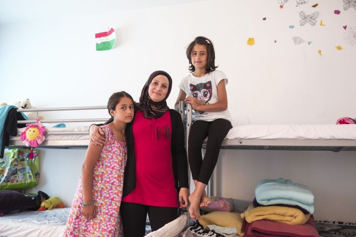 Eine Frau mit Kopftuch steht mit ihren beiden Töchtern Kindern vor einem Hochbett und lächelt in die Kamera
