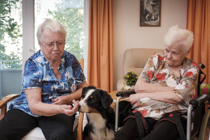Zwei ältere Frauen sitzen auf Stühlen nebeneinander. Ein Hund befindet sich zwischen ihnen und beschnuppert die Hand der Frau links im Bild.