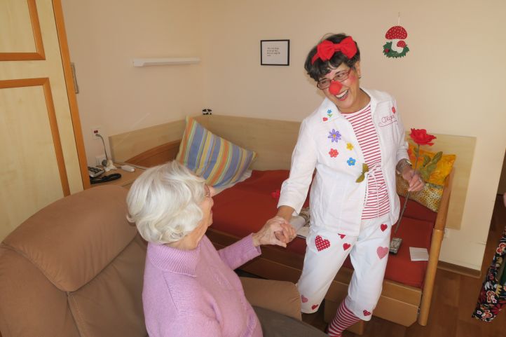 Eine Frau als ehrenamtlicher Pflegeclown tanzt mit einer sitzenden Seniorin